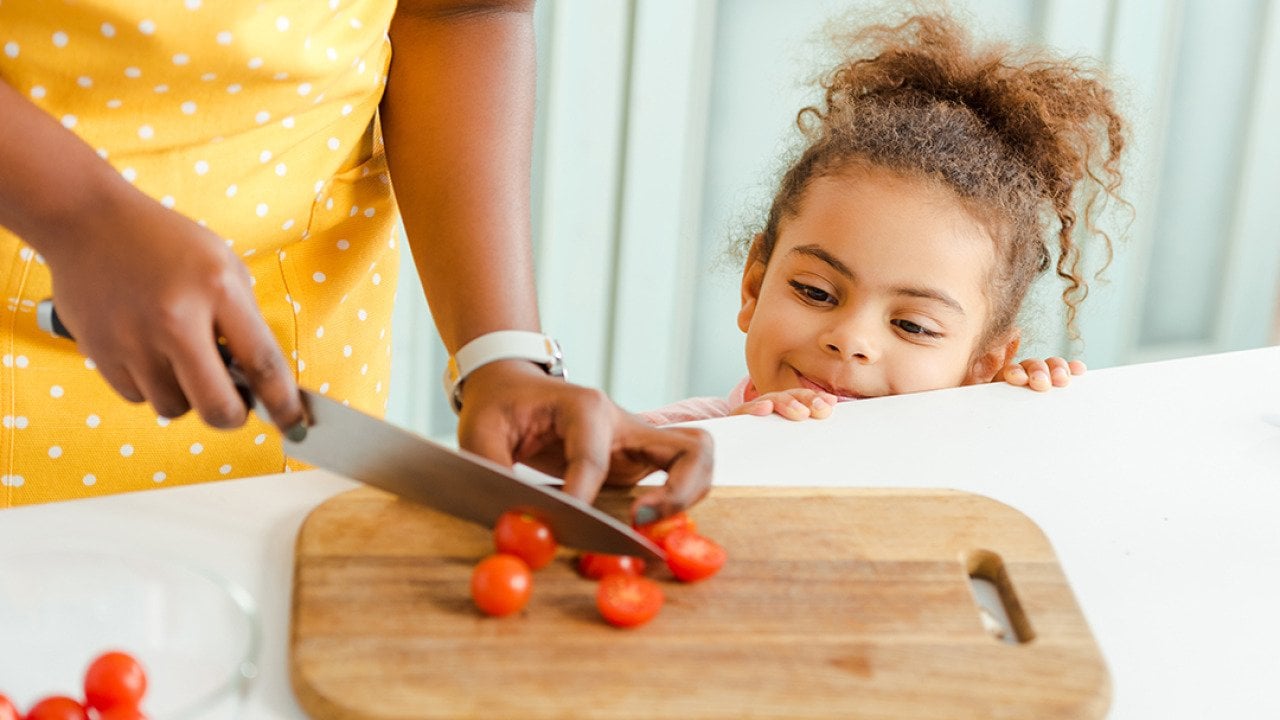 A little child watching her mother slice cherry tomatoes on a chopping board in the kitchen.