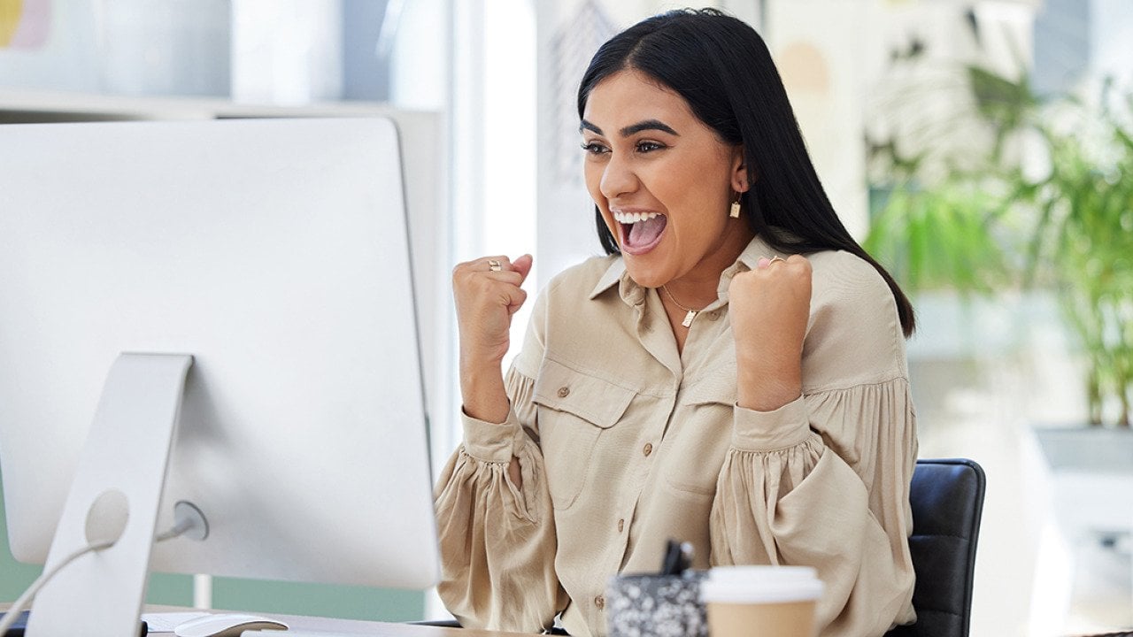 An enthusiastic businesswoman celebrates in front of her computer monitor at work.