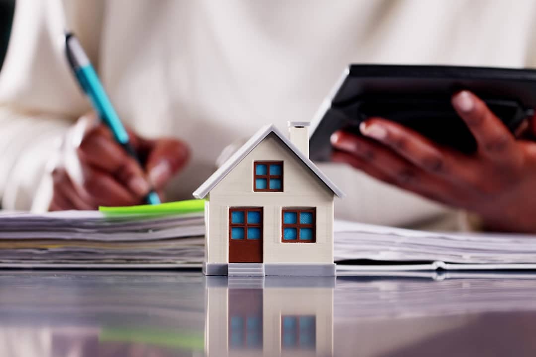 A small figure of a home with a real estate professional calculating finances in the background.