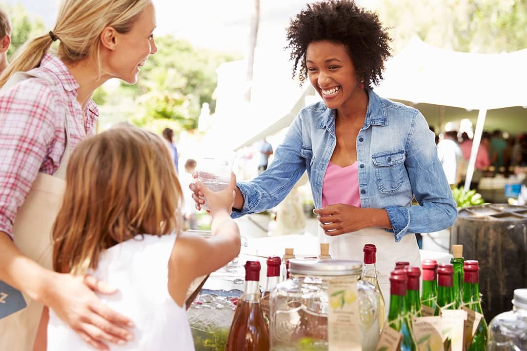 A young businesswoman selling drinks to a mother and daughter at a farmer's market stall.