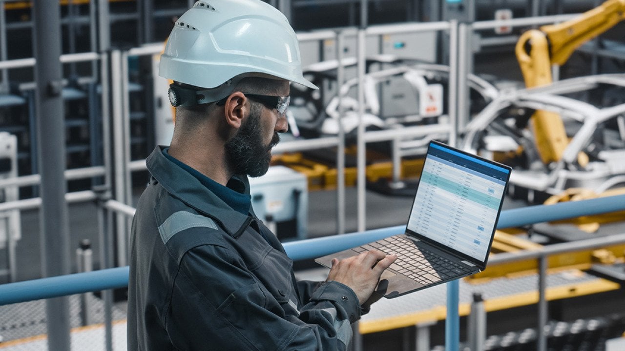 An industrial engineer working on a vehicle production spreadsheet from his laptop at a manufacturing facility.