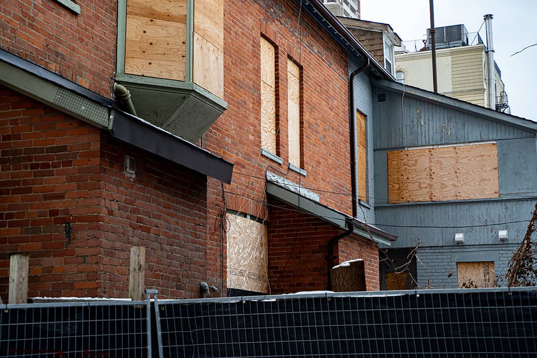 Abandoned brick and wooden houses with boarded windows enclosed by a construction fence.