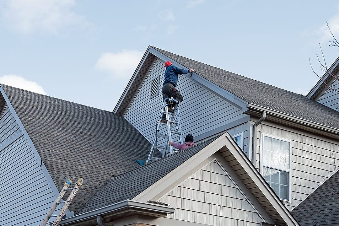 Professional roofers repairing damaged siding trim along multiple roof lines after storm damage.
