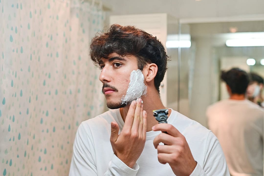 A young man shaving his beard in front of a mirror.