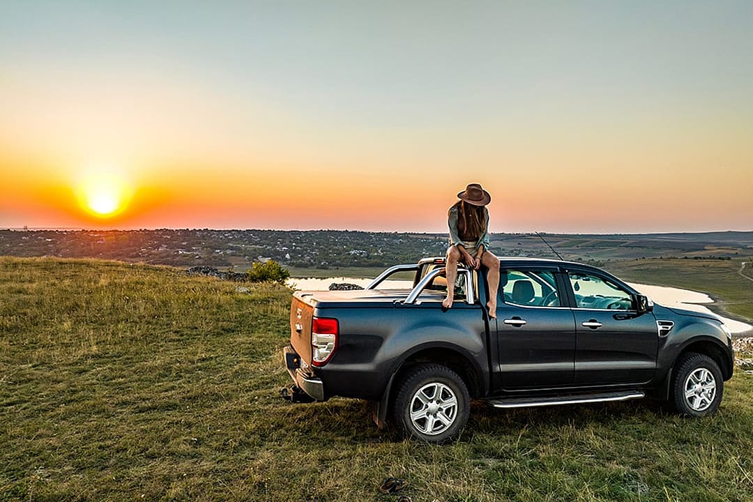 A person sitting on the roof of a pickup truck parked on a grassy hill during sunset. 
