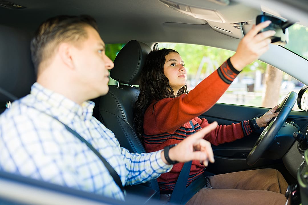 A teenage girl adjusting rearview mirror while learning to drive with her father.