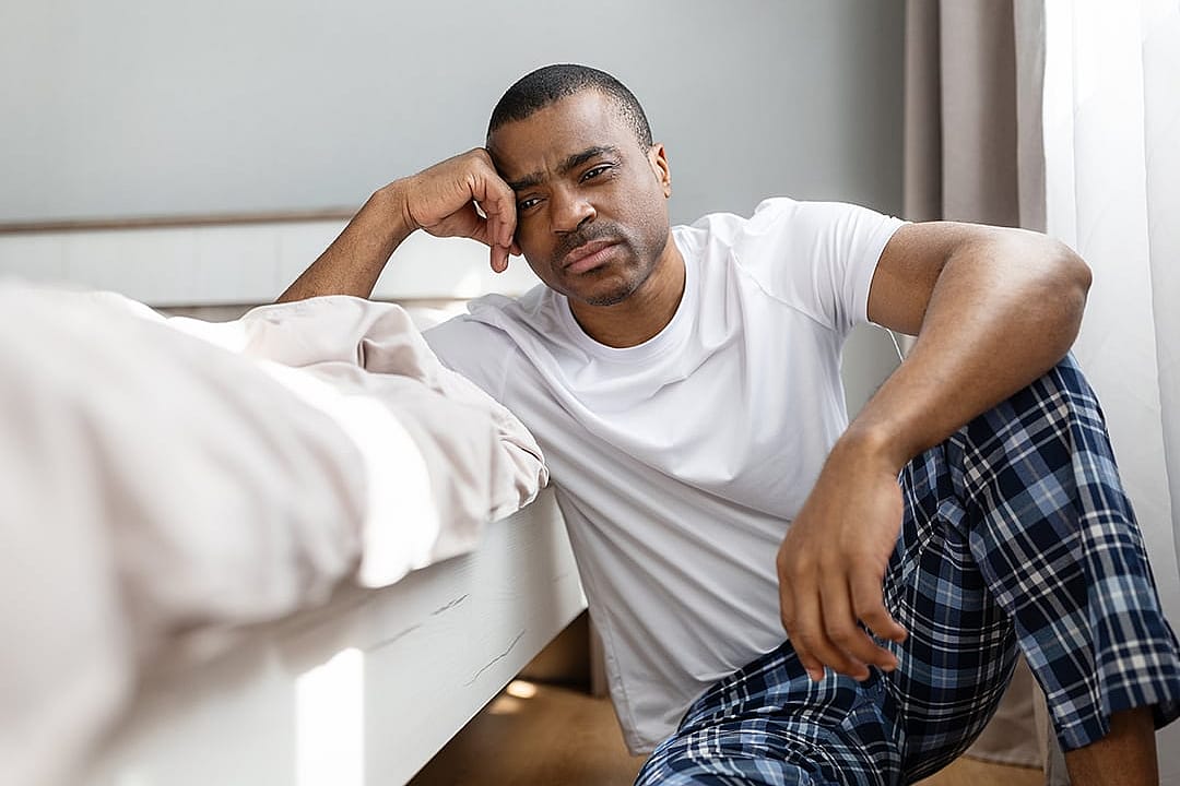 A man sitting by the end of his bed with a tired expression.