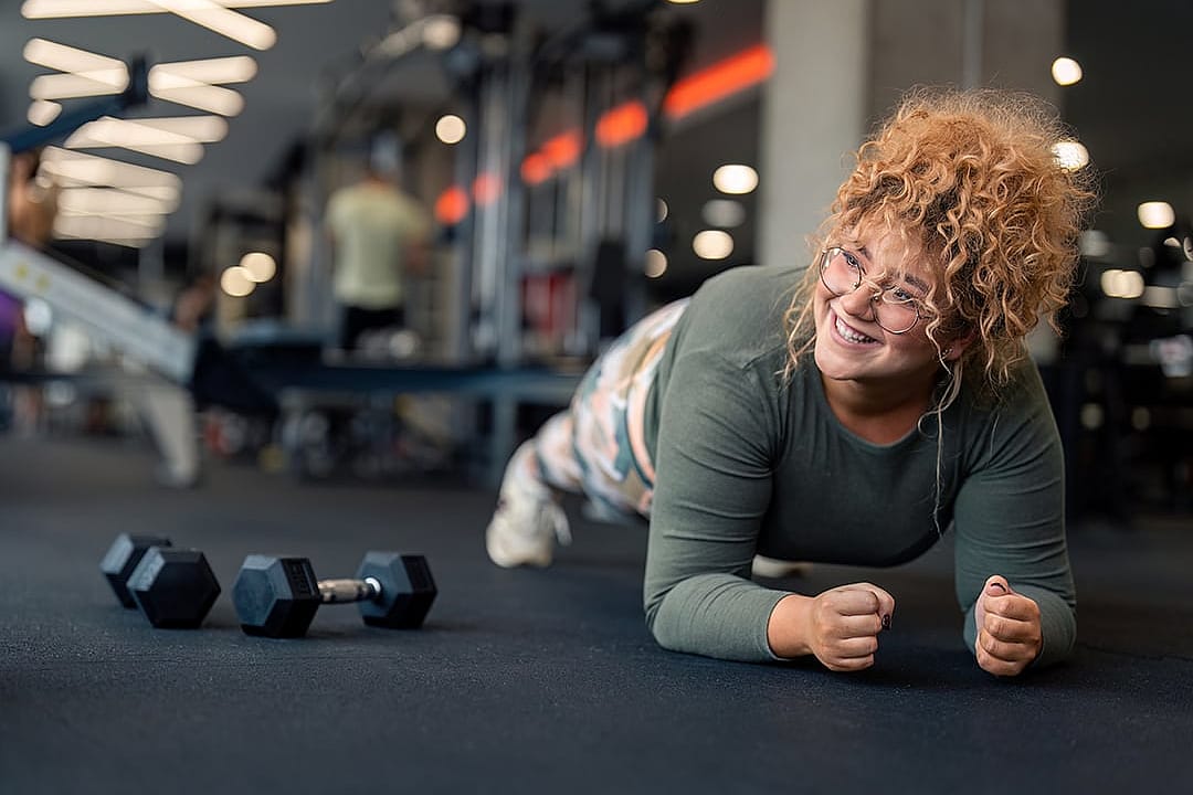A young woman performing a forearm plank exercise on a gym floor.
