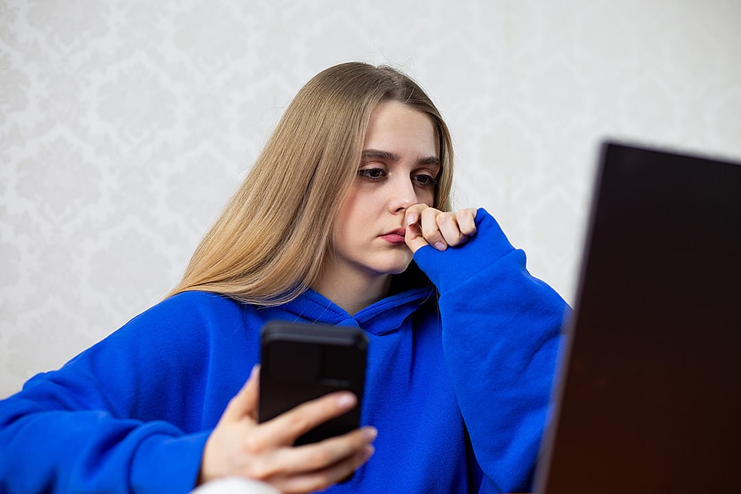 A young woman sits frozen in front of her laptop while holding her phone.