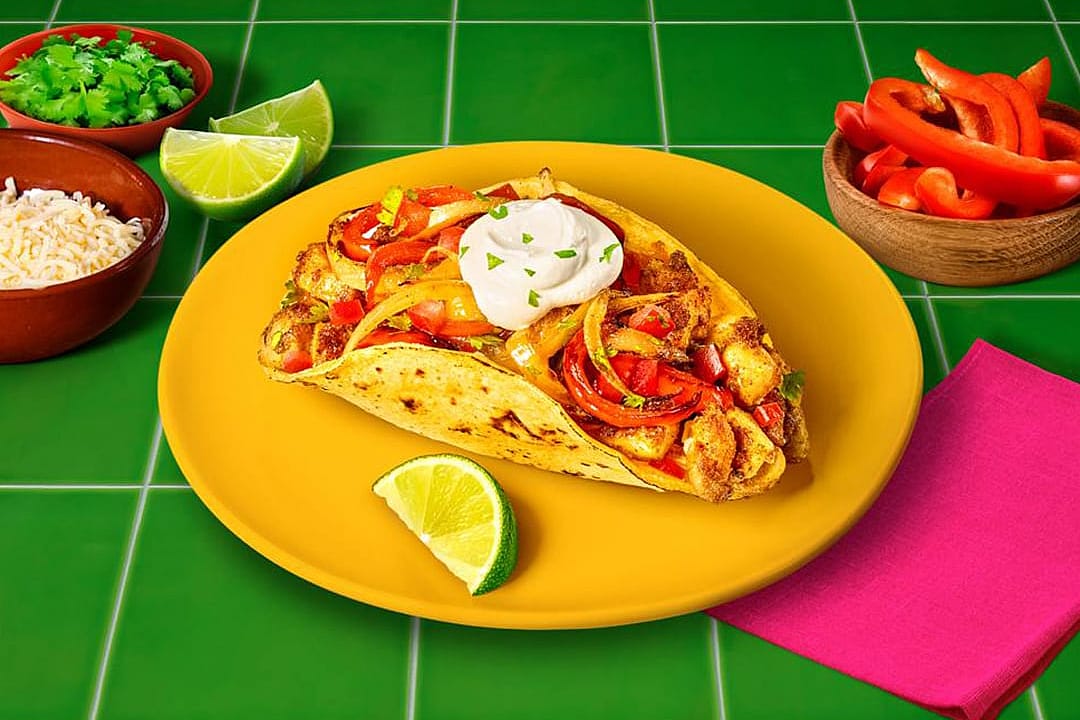 A fajita topped with sour cream served in a plate with lemon wedges, grated cheese, cilantro leaves, and bell peppers on the sides.