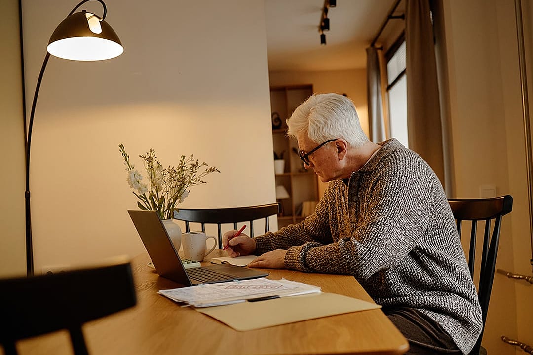 An elderly person writing notes, calculating bills, and using a laptop at home.