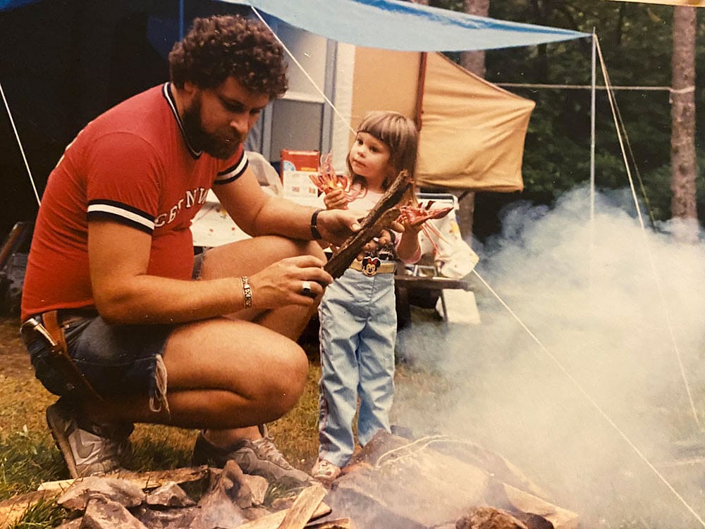 A bearded man crouches by a smoky campfire with a small girl in jeans and a Mickey Mouse belt, both holding sticks over the flames, with a tent and tarp visible behind them in the woods.