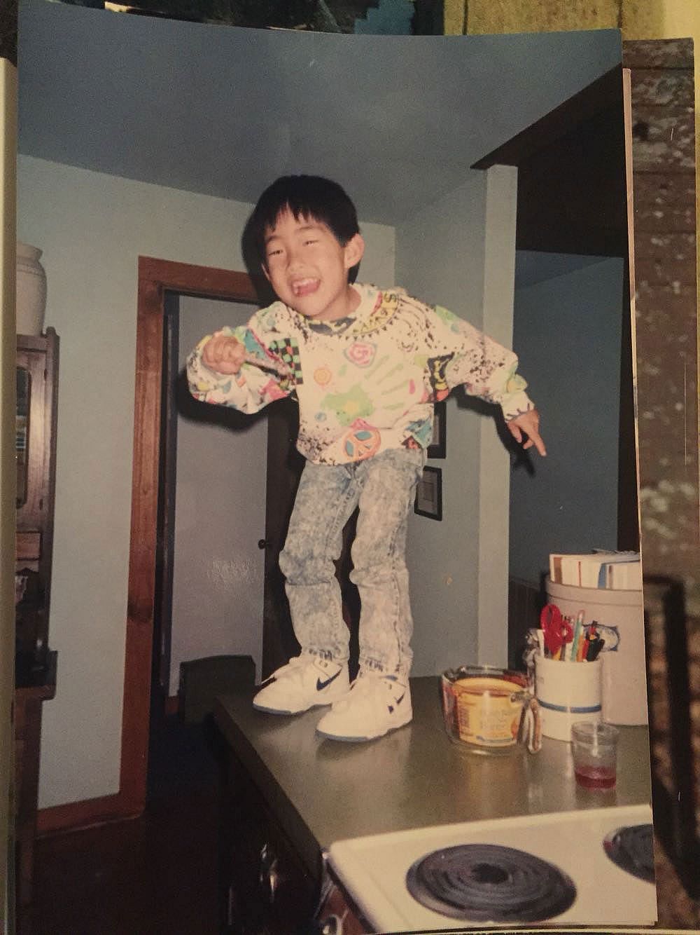 A young boy in a colorful graphic sweatshirt, acid wash jeans, and Nike high-tops dances on a kitchen counter, grinning, with a stove visible in the foreground.