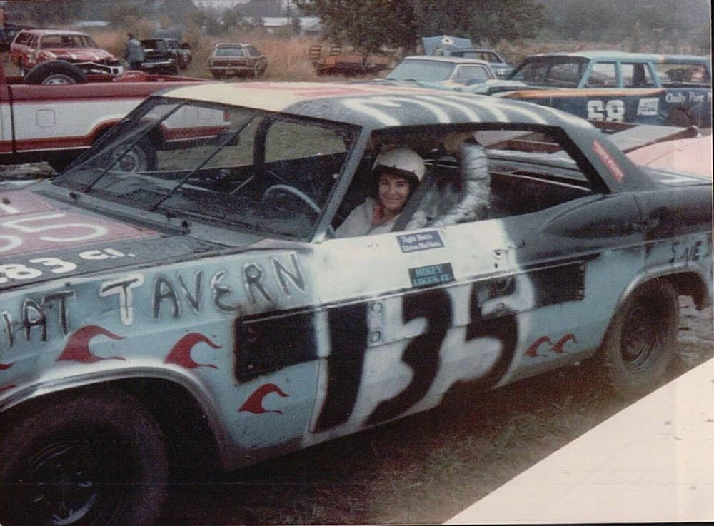 A smiling woman behind the wheel of a demolition derby car