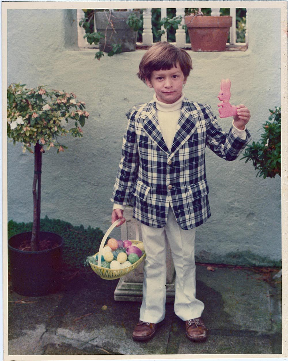 Color photo of a young boy standing in a garden courtyard in California on Easter, 1977. He wears a plaid blazer over a white turtleneck with white trousers and brown shoes, holds a yellow Easter basket full of colored eggs in one hand, and raises a pink sugar bunny cookie in the other. Potted topiary trees and a stucco wall are visible behind him.
