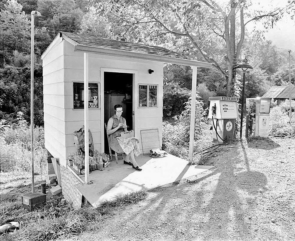 Black-and-white photo of a woman seated in the open doorway of a small white clapboard gas station in Knott County, Kentucky, in 1977. She sews in her lap while two vintage gas pumps stand to her right on a dirt lot surrounded by trees and overgrowth.