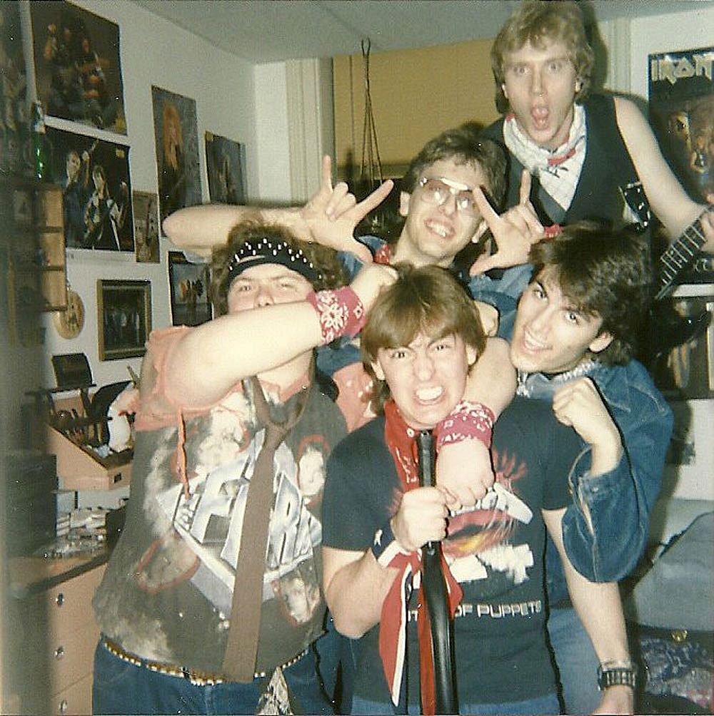 ive teenage boys crowd together in a bedroom covered in rock band posters, throwing up devil horns and mugging for the camera in band shirts, bandanas, and denim before a concert in 1987.
