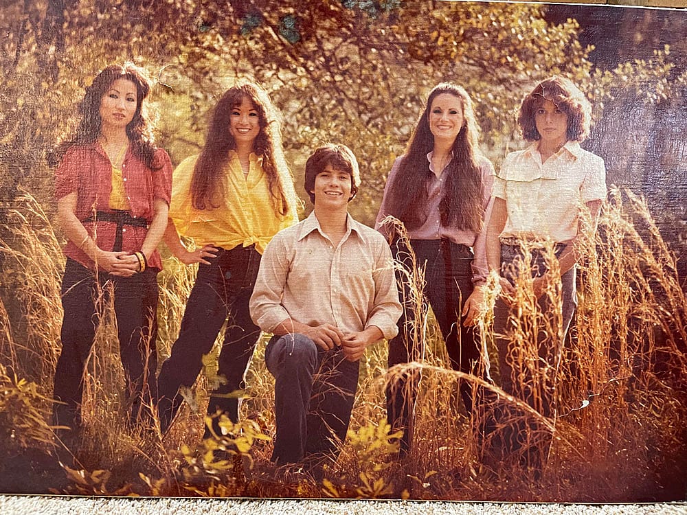 Color photo of five young siblings posing together in a field of tall golden grass in 1977. Four women and one young man stand together among autumn foliage. They wear earth-toned blouses and dark pants; several have long dark hair.