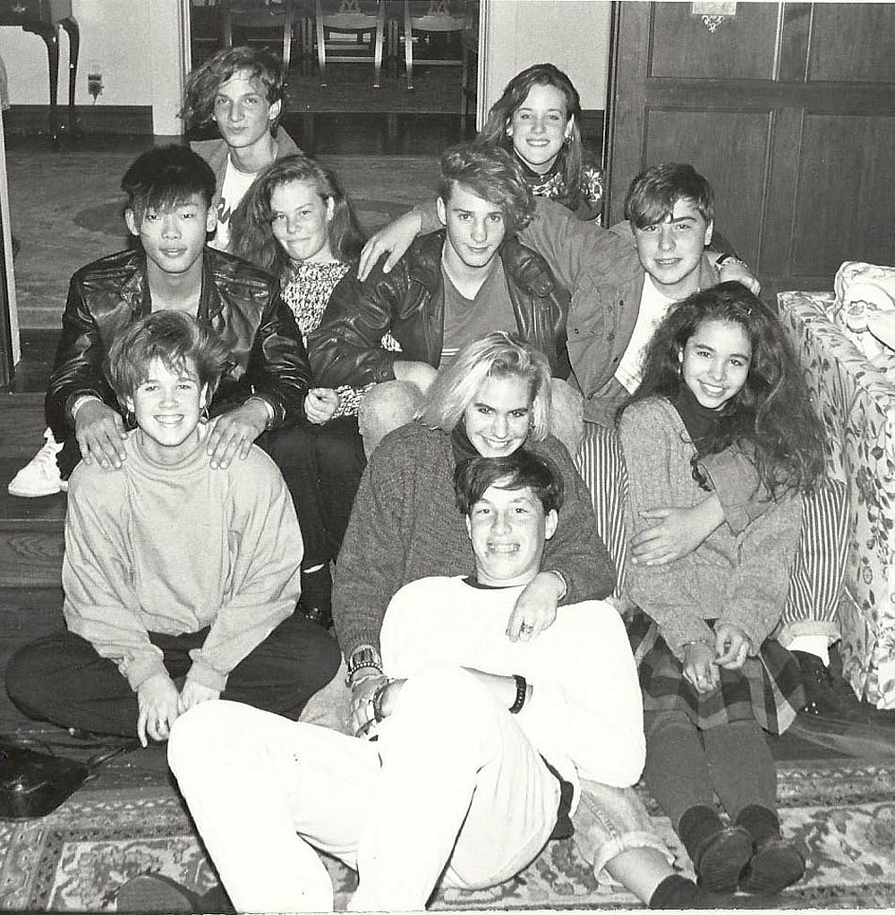 A group of about ten teenagers crowd together on the floor and a couch in a living room, smiling at the camera in a black and white photo.