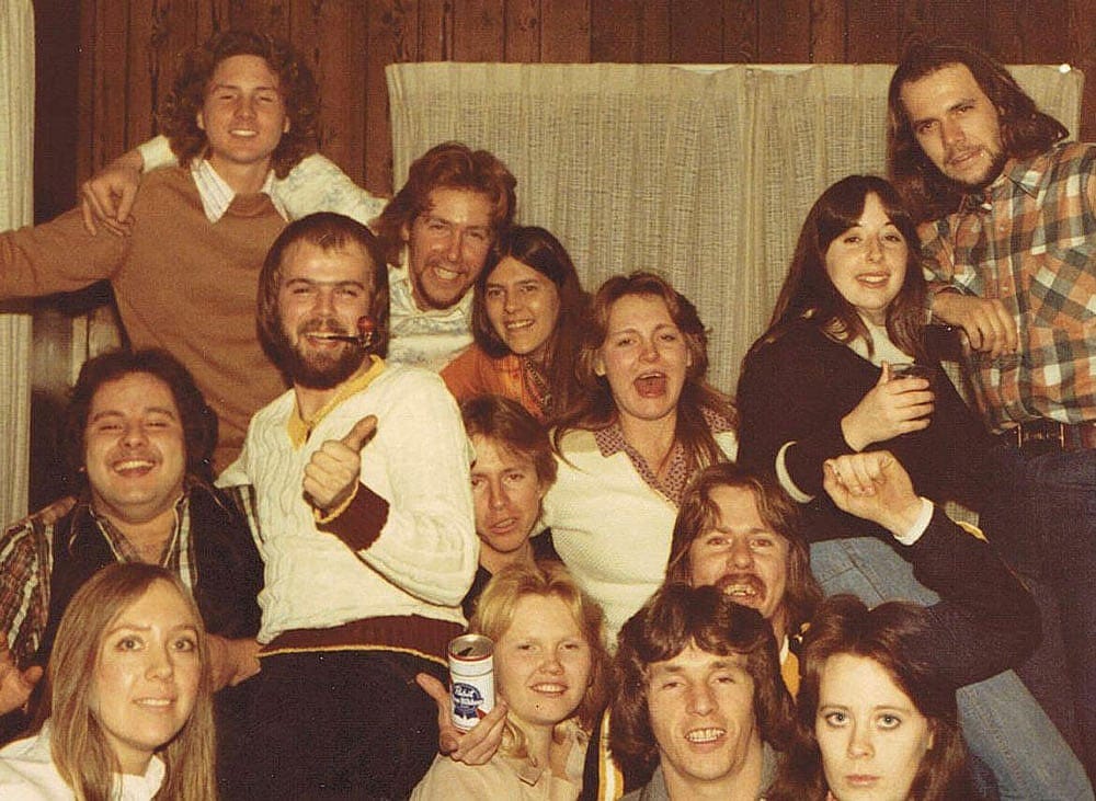 Color photo of a large group of young adults packed together in a wood-paneled living room at a birthday party in 1977. About fourteen people fill the frame, grinning and mugging for the camera. One man in a white cable-knit sweater gives a thumbs up while holding a pipe; a woman in the front holds a can of Pabst Blue Ribbon.