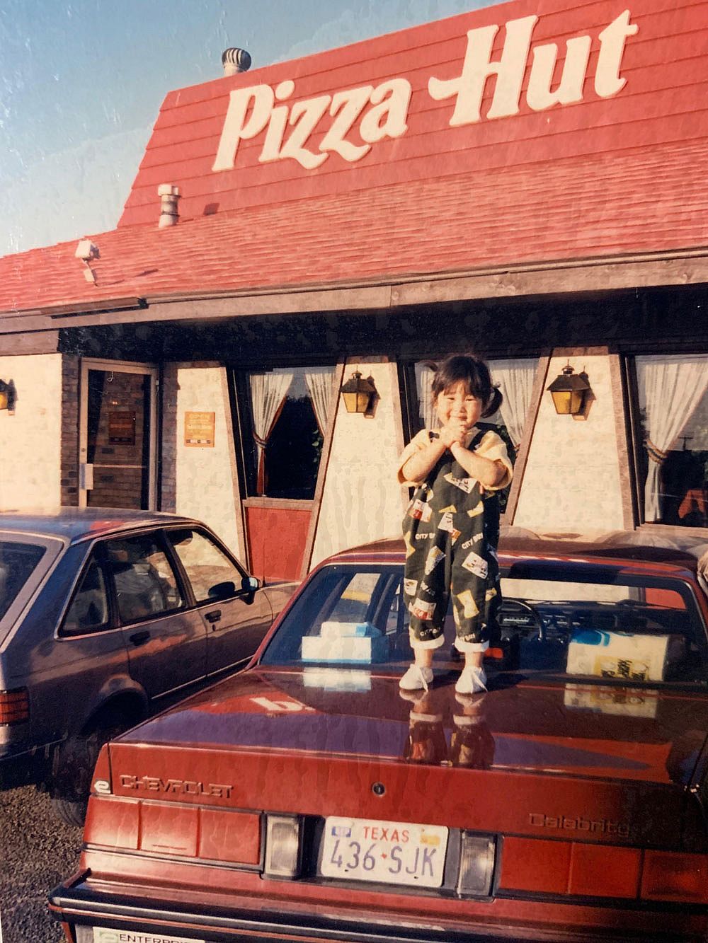 A toddler in printed overalls stands on the trunk of a red Chevrolet Celebrity in a parking lot, grinning in front of a classic red-roofed Pizza Hut building in Texas.