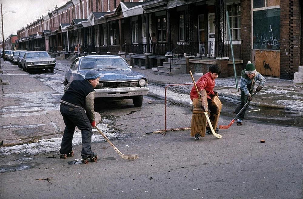Color photo of three children playing street hockey on a snowy, wet Philadelphia street in 1977. One child wearing roller skates winds up for a shot while another crouches as goalie behind a makeshift net using a wooden chair. A third player defends. A row of brick rowhouses and parked cars lines the street behind them.
