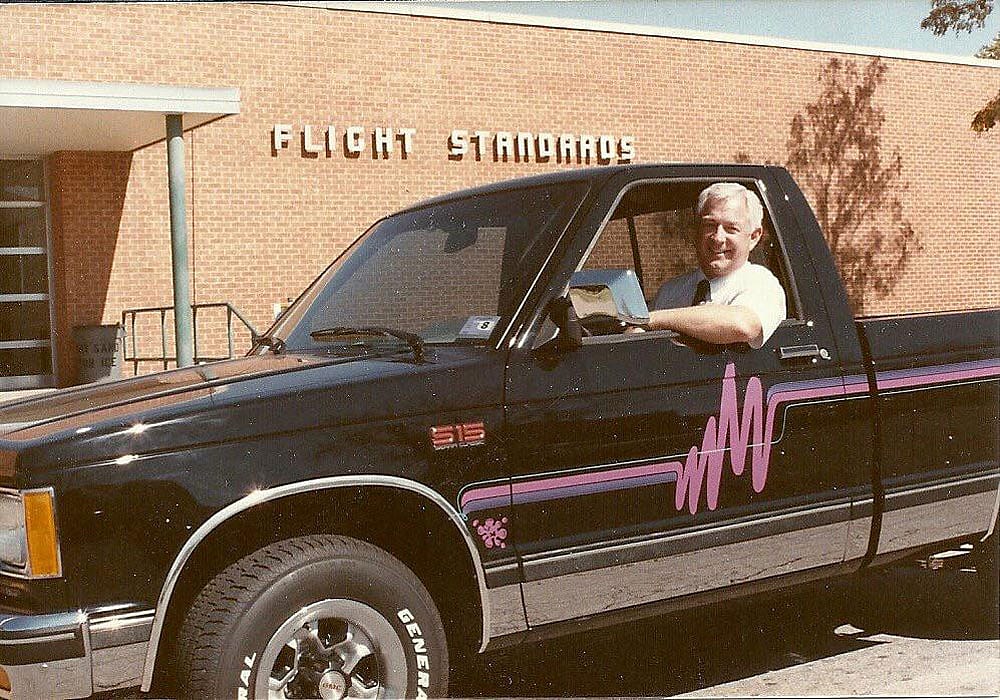 A man sits in the driver's seat of a black GMC S-15 pickup truck with bold pink and purple wave graphic decals along the side, parked in front of a brick building with a "Flight Standards" sign.