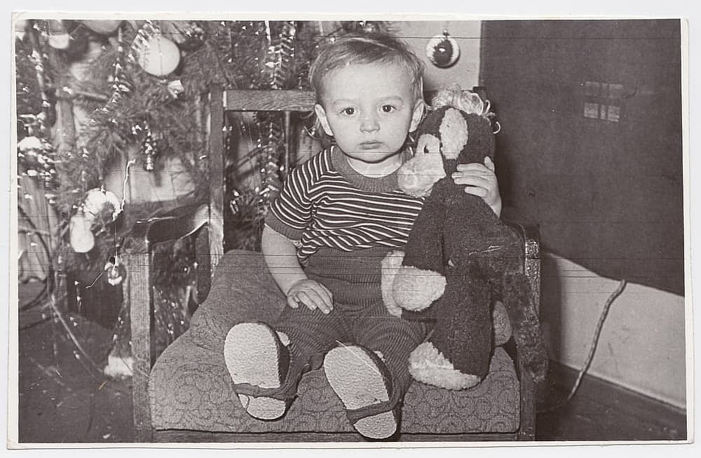 A child sits on a chair with a toy under a Christmas tree in an old black-and-white photograph.