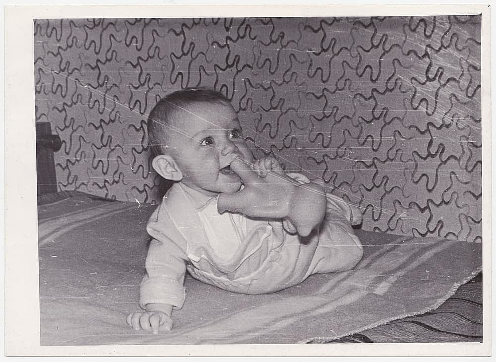 A vintage photograph of a beautiful baby crawling on a crib, holding a toy to his mouth. The photograph captures the warm atmosphere of yesteryear.