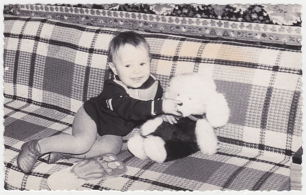 An old sweet photo of a cute cheerful boy sitting on a sofa with a toy. Vintage black and white photo, slightly yellowed by time, taken in the USSR around the 1960s.
