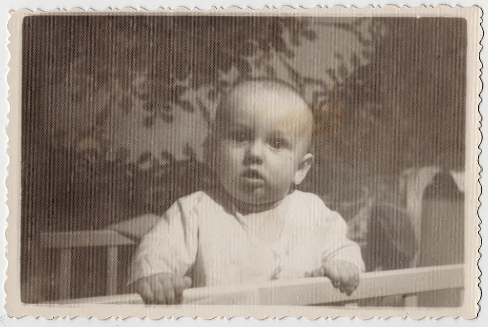 Vintage photo of a baby in a crib in an apartment. An old, not very clear, grainy black and white photo, slightly yellowed with age. The photo was taken in the USSR around the 1960s.