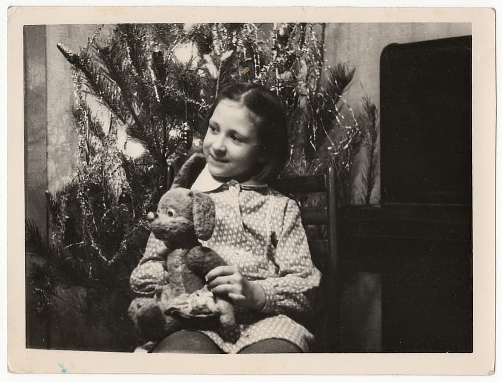A girl sits on a chair and holds a teddy bear in her hands near a Christmas tree in an apartment in a vintage black and white photograph. The old photo was taken in the USSR in the 1960s.