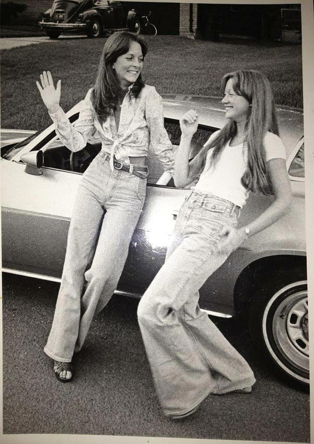 Black-and-white photo of two young women leaning against a car in a suburban driveway in 1977