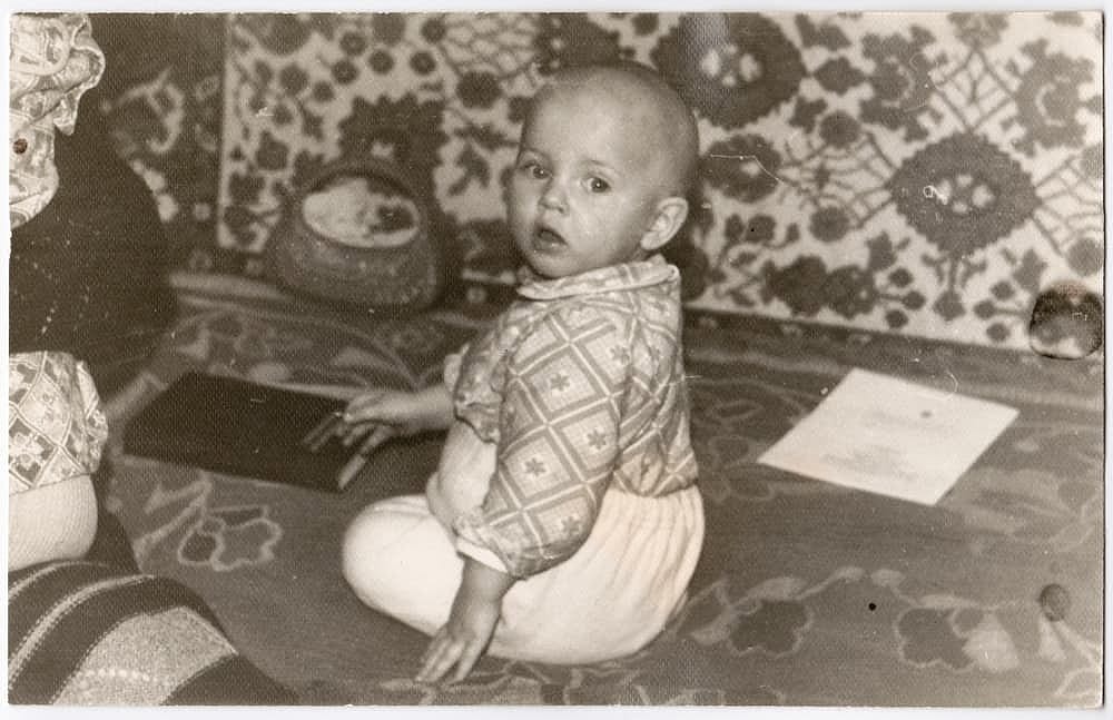 An old cute photo of a baby in an apartment. Vintage black and white textured photograph, slightly yellowed with age, taken in the USSR around the 1960s