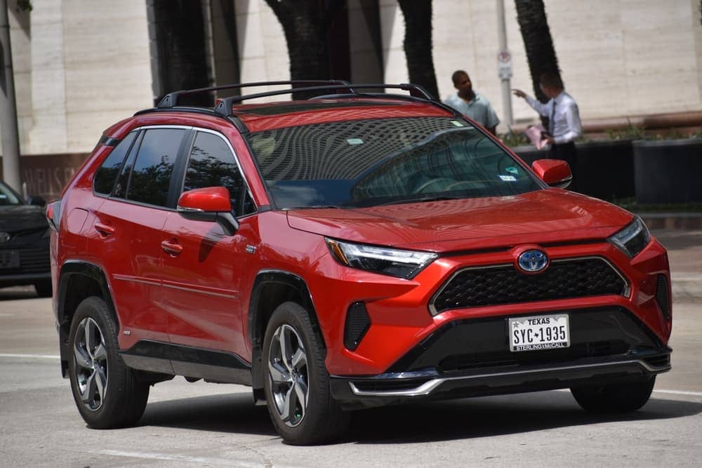 A portrait of a red Toyota RAV4 Prime SUV cruising in a downtown financial district.