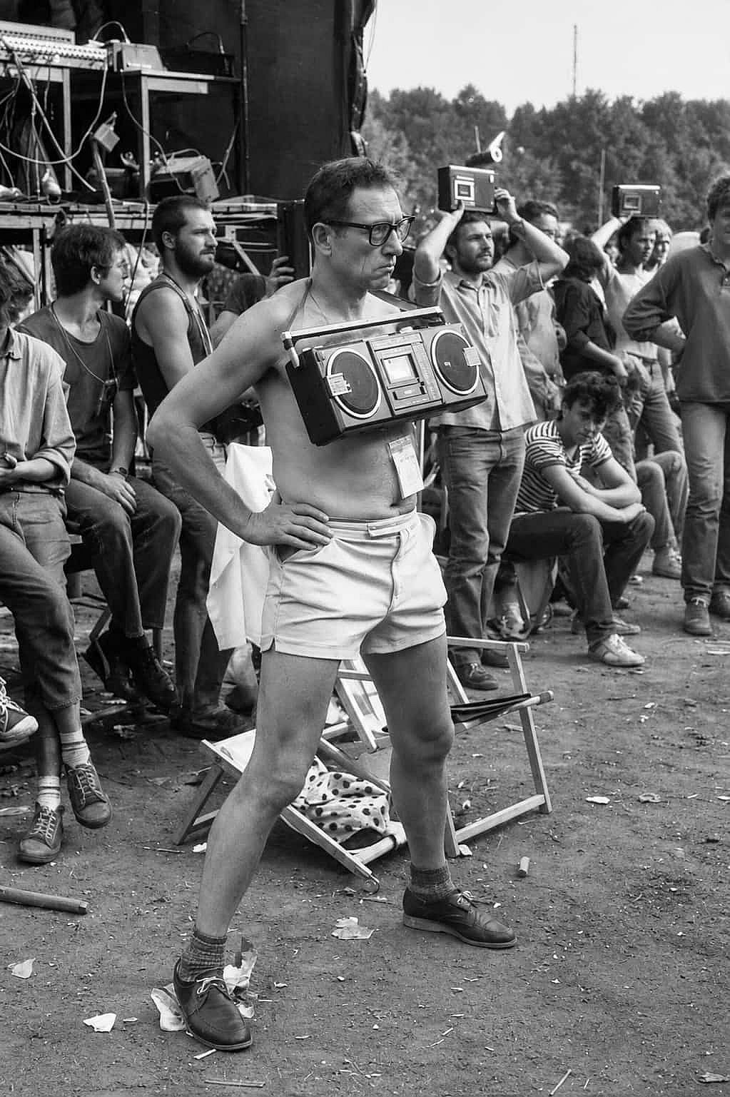 A man with a boombox hanging from his neck is recording a concert in 1980.