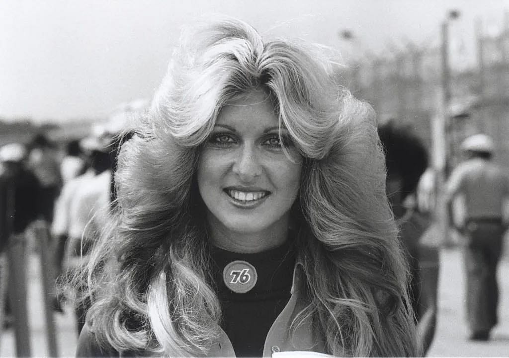 Black-and-white close-up photo of a woman with large, voluminous feathered blond hair at Daytona Bike Week, circa 1977. She wears a Union 76 pin on her collar and smiles at the camera. A crowd of people is visible in the soft-focus background.