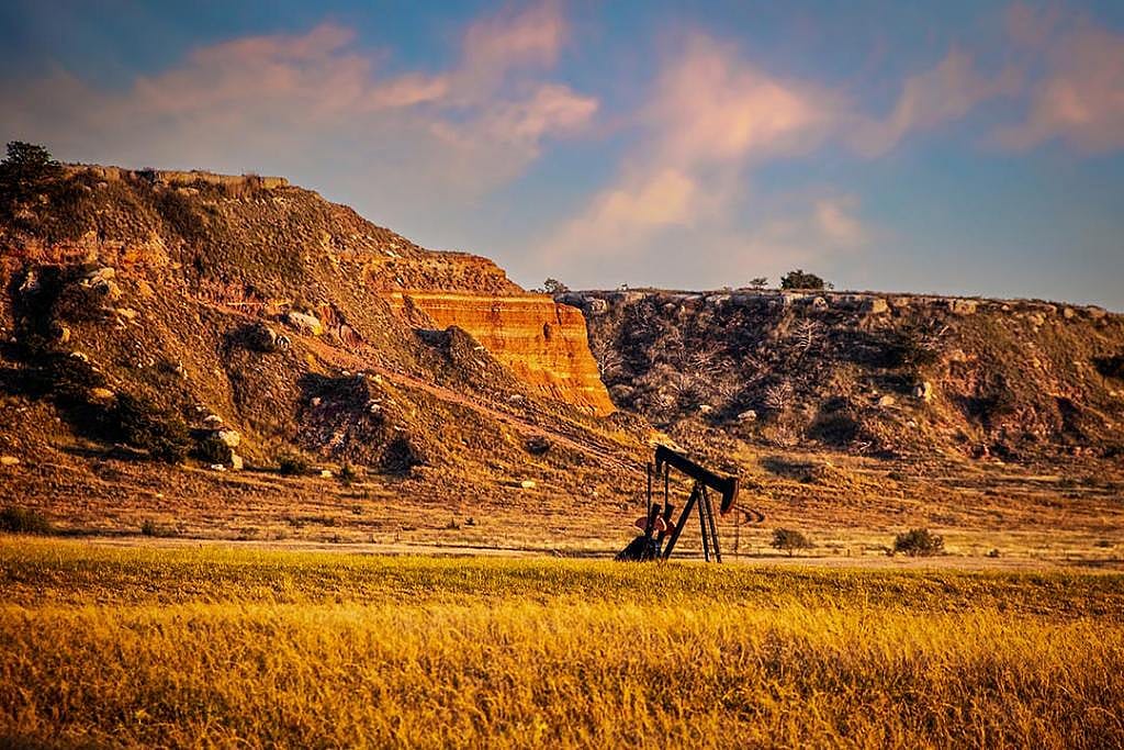 Oil well pump jack in red earth western Oklahoma at golden hour with early sunset sky and bluffs in background.