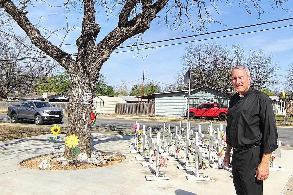 Father Michael Marsh stands in front of a memorial for the Uvalde shooting victims which is on the ground next to a tree with no leaves.