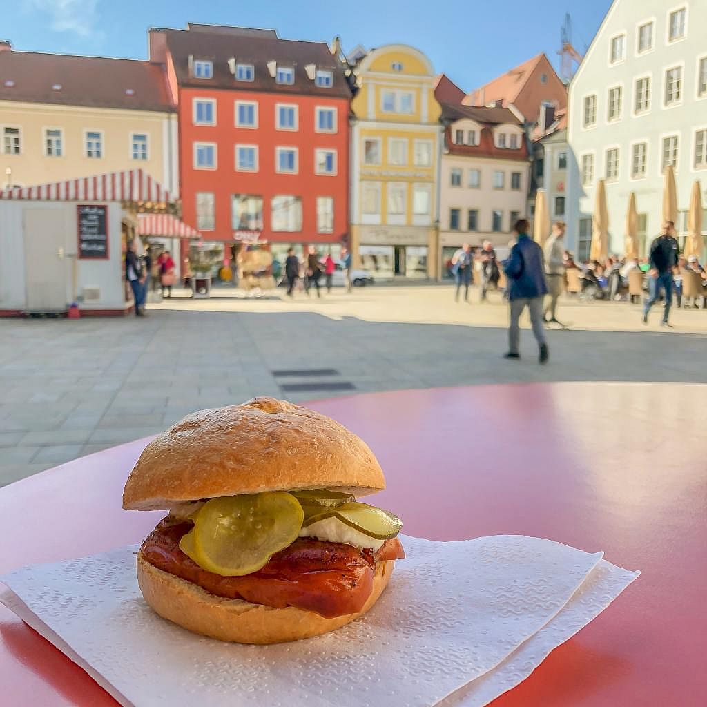 A Knacker, a one-of-a-kind Bavarian sausage served on a bun on a napkin on a red table in front of picturesque German buildings along the Danube River in eastern Bavaria.