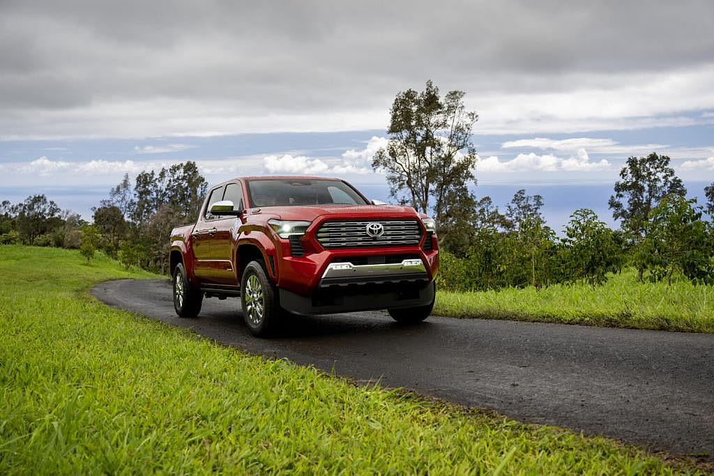 Front view of red 2026 Toyota Tacoma driving on an asphalt road with green grass on either side and gray clouds in the sky.