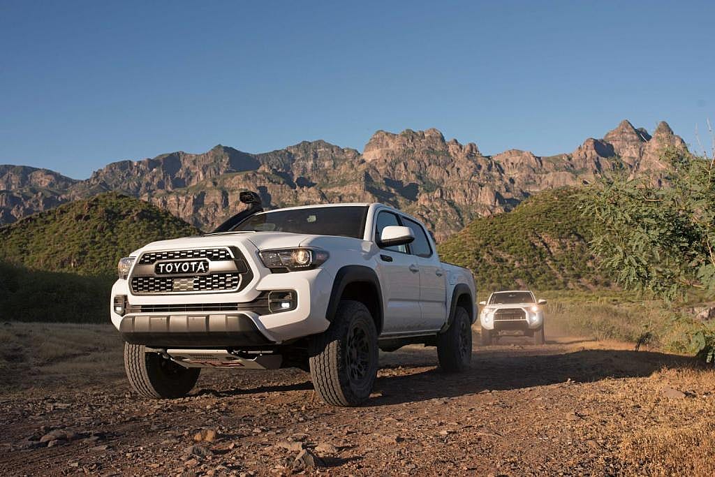Front view of white 2019 Toyota Tacoma driving on dirt road with rugged mountains in the background. Another white truck with headlights on can be seen following behind.