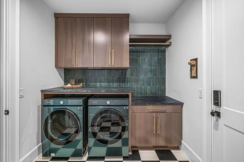 Modern laundry room with dark turquoise appliances, tiled backsplash, wooden cabinets and with brass hardware and a black-and-white checkerboard floor.