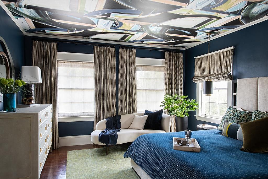 Bedroom with large windows, navy blue walls and a design painted on the ceiling and furnished with bed, dresser, settee, and matching decor like lamps, rugs, pillows and plants.