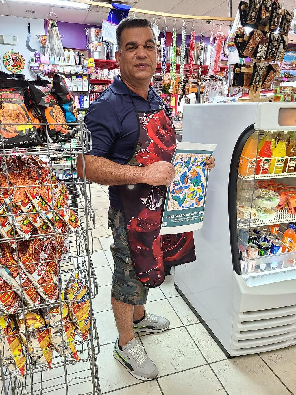 Man wearing a red floral apron holds a poster in a small standing between snack and drink displays. Resources for 4th Amendment Workplaces include a workplace guide, organizing toolkit, posters signaling opposition to unconstitutional search and seizures, employee handouts and tips for designating private employee areas.
