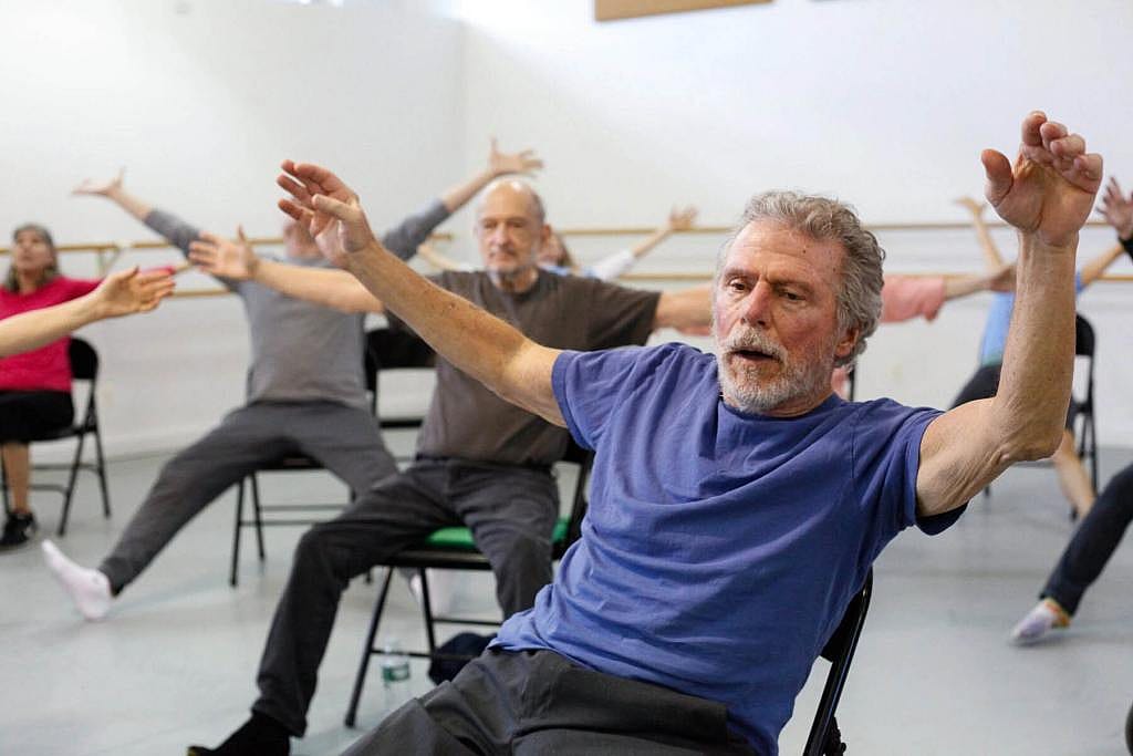Elderly male Dance for PD participants extend their arms and legs while sitting on a chair.