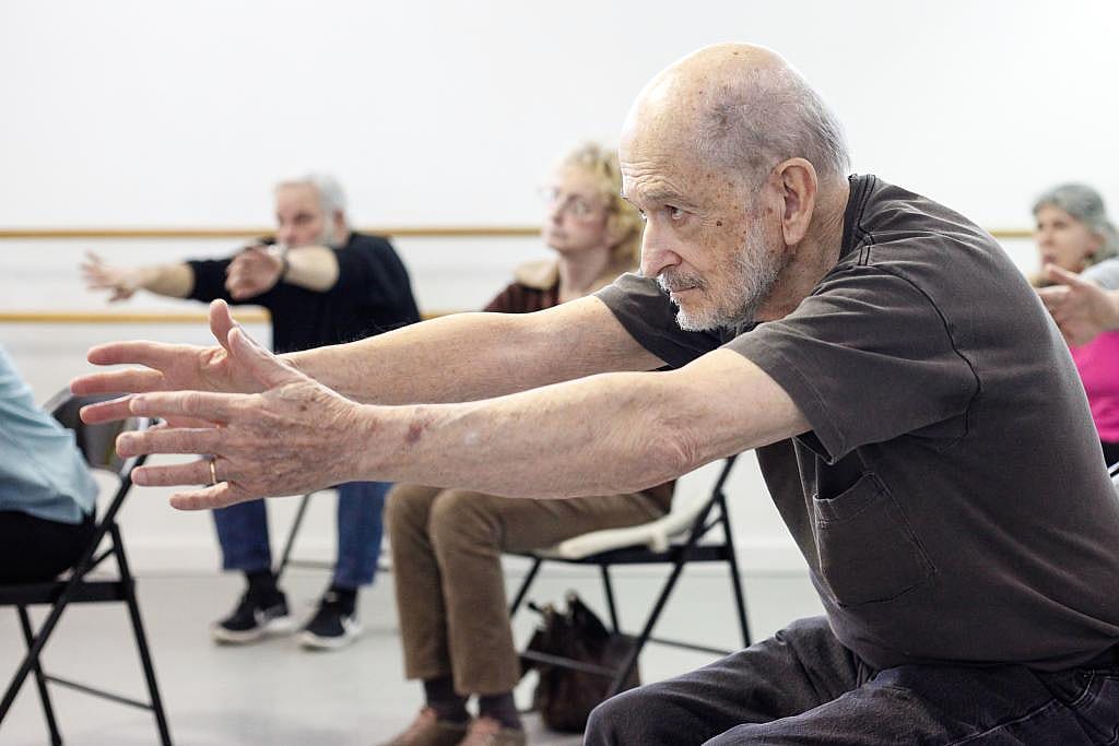 A man wearing a brown shirt extends his arms forward in a pose while sitting on a chair as part of a Dance for PD program.