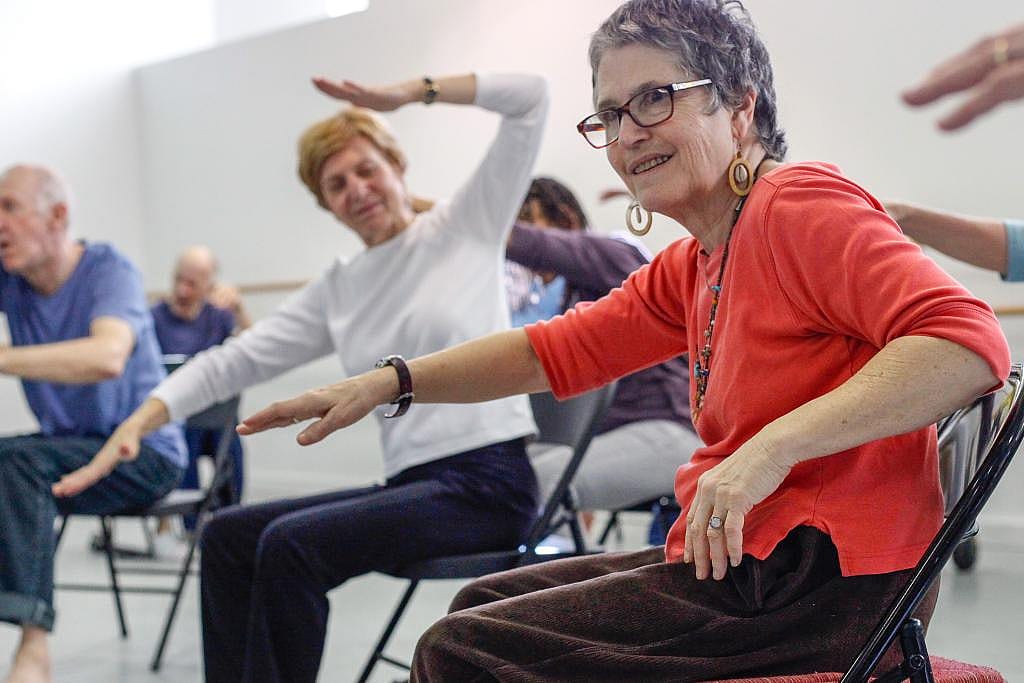 A woman in the Dance for PD program wearing an orange shirt dances from a chair in the foreground, while others are visible behind her doing the same.