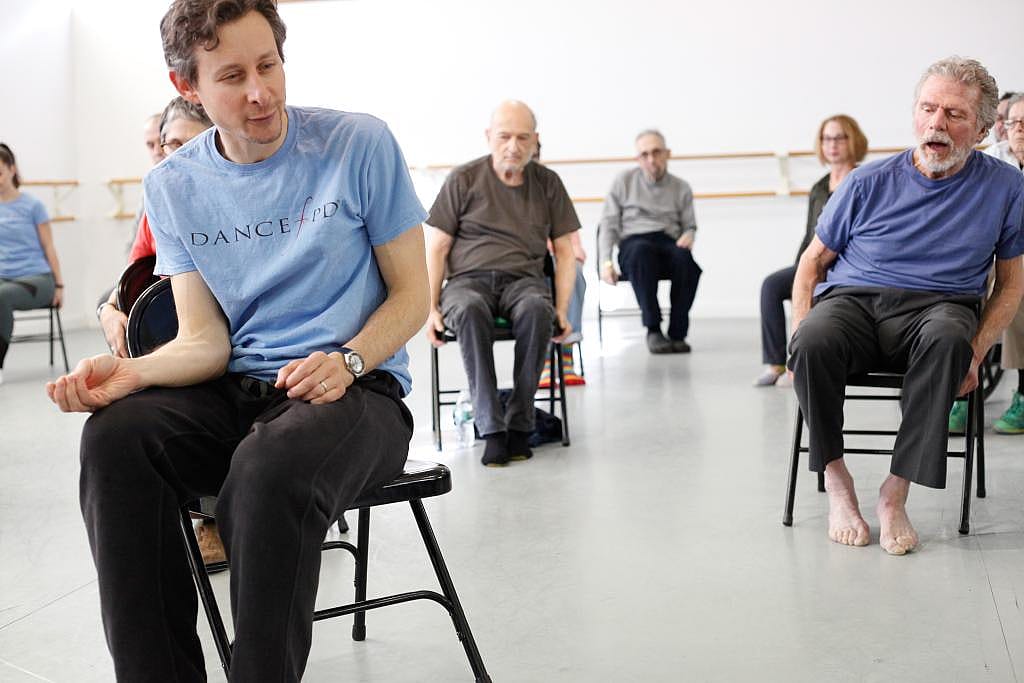 A person wearing a light blue shirt in the foreground with many people in the background all doing chair dance.
