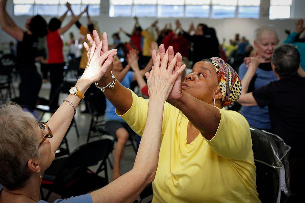 A woman wearing a yellow shirt with raised hands dances palm-to-palm with a person sitting across from her in a Dance for PD program.